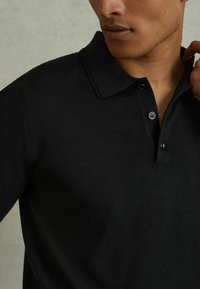 Svart polo skjorta tillverkad av ett slätt, strukturerat tyg; har en krage, knappslå och en figurnära design.