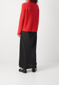 Rode geribde coltrui, gecombineerd met een zwarte maxi rok en platte zwarte schoenen. Gladde textuur met aansluitende mouwen.