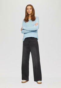 Helles, blaues Langarm-Pullover mit lockerer Passform, kombiniert mit schwarzen weit geschnittenen Jeans. Das Model trägt legere, beige Schuhe mit glanzender Oberfläche.