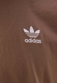 Hnědé bavlněné tričko s vyšitým bílým logem Adidas trefoil na hrudi. Textura je hladká s jemným žebrovaným vzorem.