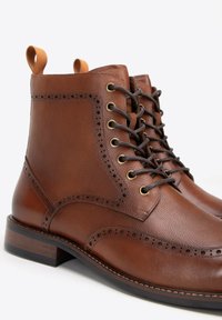 Hnědé kožené kotníkové boty s brogue detaily, zlatými očičky, hnědými tkaničkami a zvýšenou patou. Obsahují poutko na límci.