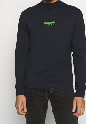 Mann in einem schwarzen Langarmshirt mit einem kleinen grünen und schwarzen Calvin Klein-Logo auf der Brust, kombiniert mit dunklen Jeans.