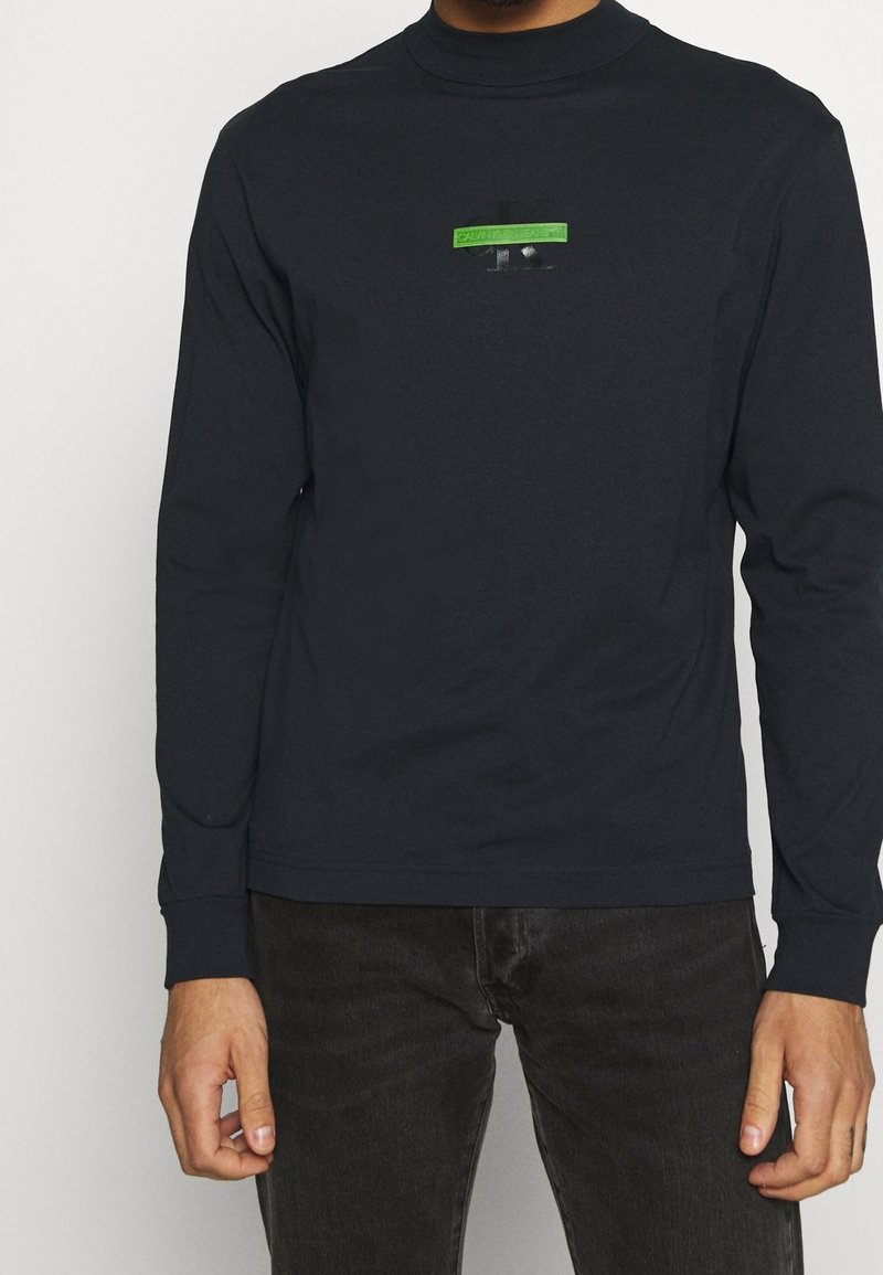 En man som bär en svart långärmad skjorta med en liten grön och svart Calvin Klein-logotyp på bröstet, tillsammans med mörka jeans.