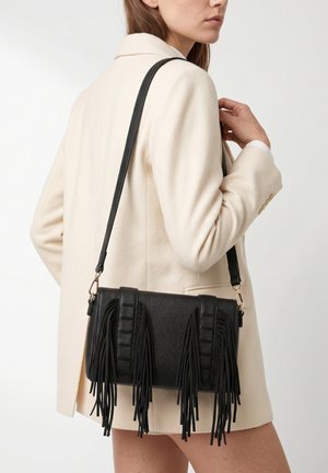 Vrouw draagt een crèmekleurige blazer en draagt een zwarte leren handtas met lange franjes over haar schouder.