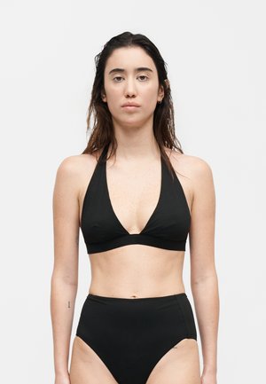 Γυναίκα με μακριά σκούρα μαλλιά, φορώντας μαύρο τοπ μπικίνι με halter και μπικίνι υψηλής μέσης, στέκεται μπροστά σε απλό φόντο.