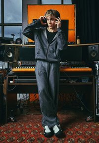 Set di felpa grigia con cappuccio e pantaloni della tuta caratterizzato da grafica ricamata, abbinato a sneakers bianche e nere. Sfondo da studio con attrezzatura audio.