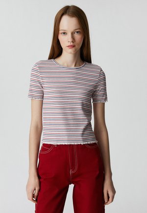 Gestreepte top met korte mouwen in het wit met rode en blauwe lijnen, voorzien van een geribbelde textuur en een cropped zoom. Gecombineerd met rode broeken.