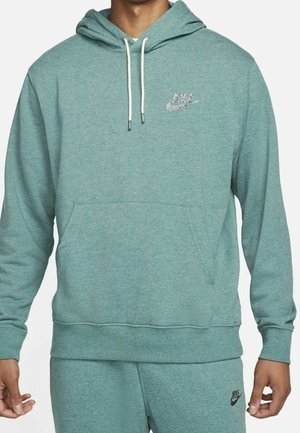 Man draagt een teal Nike-hoodie en bijpassende broek met witte trekkoorden en zwarte Nike-logo's op de borst en de dij.