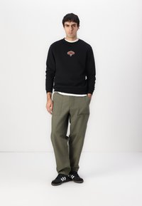 Black sweatshirt with logo, white crew neck shirt underneath, olive green wide-leg trousers, and black sneakers with white stripes.