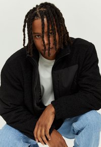 Black fleece jacket with a zip-up front, textured plush material, and a contrasting black pocket. Worn over a white shirt, paired with blue jeans.