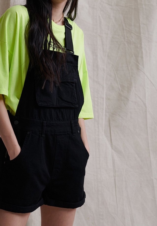 Personne portant un t-shirt oversize jaune néon avec une salopette courte en denim noir, mains dans les poches, sur un fond clair en tissu uni.