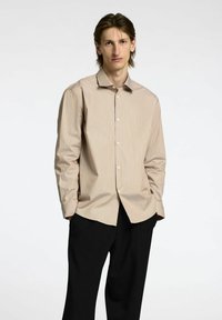 Camicia a maniche lunghe, con bottoni, di colore beige chiaro con sottili righe verticali. Presenta un colletto classico e una vestibilità dritta, abbinata a pantaloni scuri.