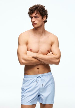 Młody mężczyzna z kręconymi włosami stoi bez koszulki, z założonymi ramionami, ubrany w jasnoniebieskie kąpielówki na jednolitym białym tle.