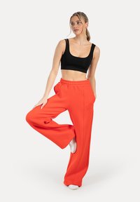 Schwarzes Crop-Top und weit geschnittene orangefarbene Hose. Die Hose hat eine hohe Taille und Falten. Das Model steht auf einem Bein und präsentiert die Hose.