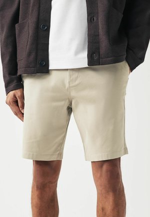 Beige shorts with a smooth texture, straight fit, and subtle stitching details, paired with a dark jacket over a white shirt.