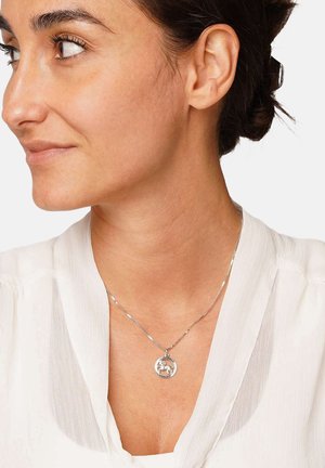 Frau mit dunklem Haar im Dutt, trägt eine weiße Bluse und eine silberne Halskette mit einem Anhänger in Form eines Baumes in einem Kreis.