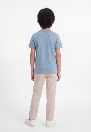 Enfant aux cheveux foncés, debout de dos, portant une chemise à manches courtes bleu clair, un pantalon beige et des baskets blanches sur fond blanc.