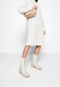 Bottes cheville beiges avec une finition texturée, talons épais et fermetures éclair sur les côtés, associées à une robe pull blanche en tricot et un sac beige.