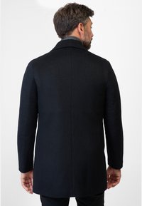 Zwarte jas met een tailormade ontwerp, gladde textuur en een subtiel visgraatpatroon. Middellange lengte met een gestructureerde kraag en enkele split.