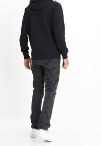 Sudadera negra con bolsillo tipo canguro, llevada sobre jeans gris oscuro y zapatillas blancas, vista desde atrás contra un fondo liso.