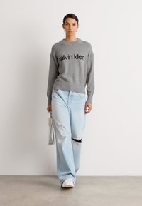 Suéter gris de punto con el logo de "Calvin Klein", combinado con jeans desgastados de color azul claro. Modelo sosteniendo un bolso plateado, de pie sobre un fondo neutro.