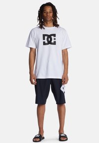 Weißes Baumwoll-T-Shirt mit schwarzem DC-Logo, kombiniert mit marineblauen Shorts mit einem weißen Akzent. Schwarze Schlappen runden das Outfit ab.