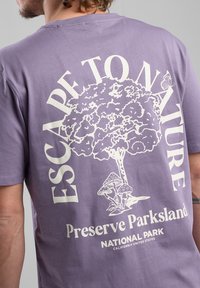 Purple cotton t-shirt featuring a white print of a tree, mushrooms, and text "Escape to Nature" and "Preserve Parkland, National Park."