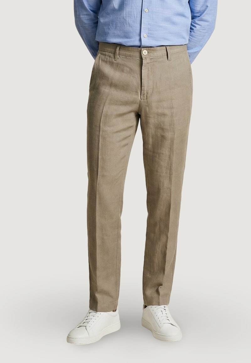 Hombre vestido con pantalones beige a medida, camisa azul claro abotonada y zapatillas blancas, de pie con las manos detrás de la espalda sobre un fondo liso.