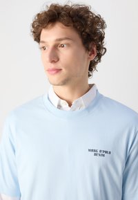 Világoskék pamut póló, kerek nyakkal, bal mellrészen fekete nyomatú "MARC O'POLO DENIM" felirattal. Egyszerű dizájn, rövid ujjak.