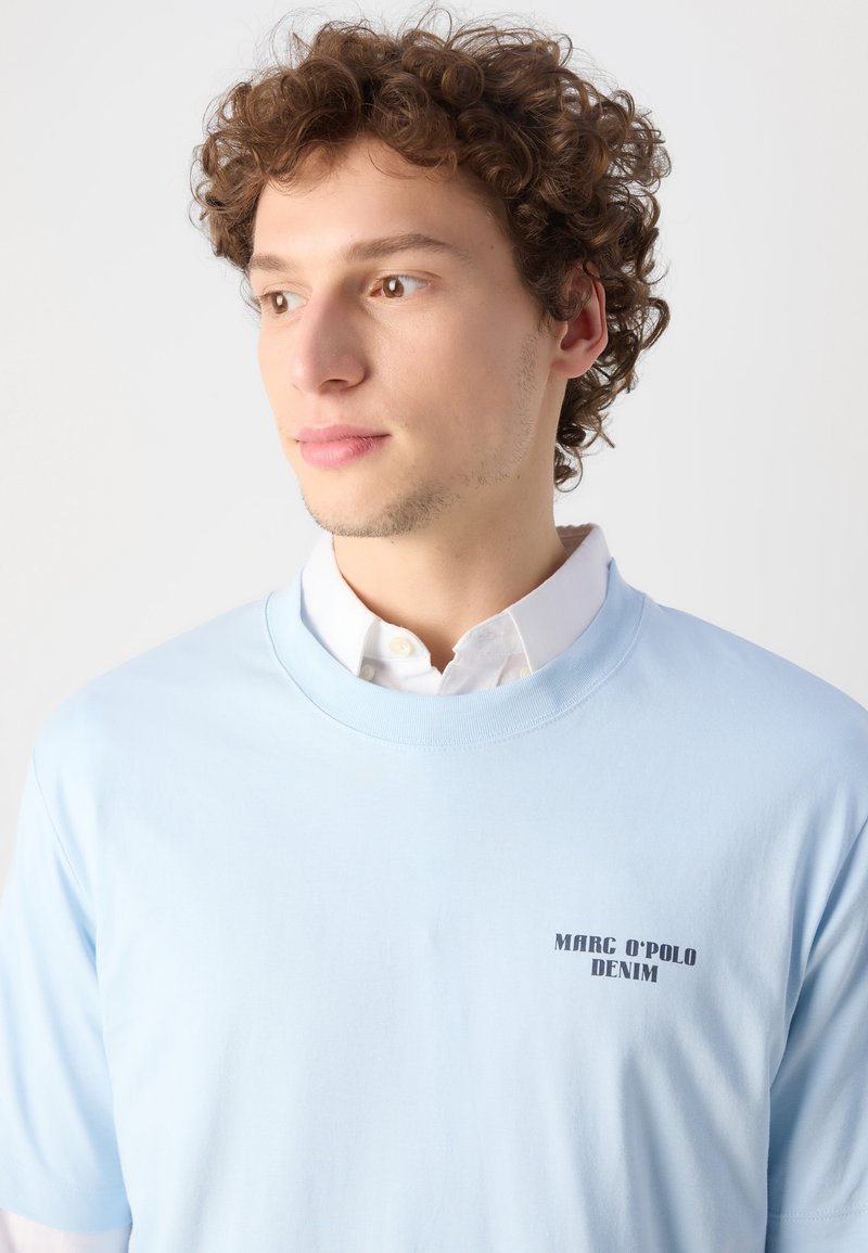 Helles, blaues Baumwoll-T-Shirt mit rundem Ausschnitt, auf der linken Brust mit einem schwarzen Aufdruck "MARC O'POLO DENIM". Einfaches Design, kurze Ärmel.
