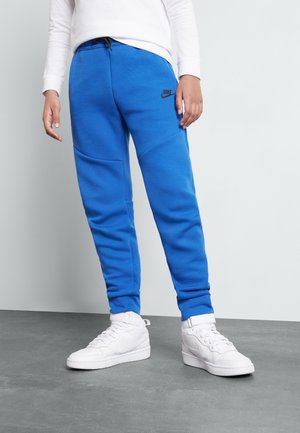 Niebieskie spodnie dresowe o gładkiej fakturze, z czarnym logo Nike, kieszeniami po bokach i ściągaczami na nogawkach. Połączone z białymi wysokimi trampkami.