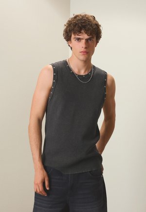 Jeune homme aux cheveux bouclés portant un haut sans manches gris foncé et un pantalon foncé, debout devant un fond clair uni.