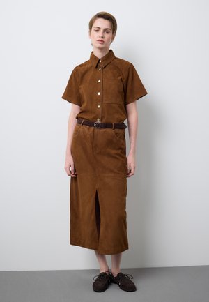 Persona con cabello corto que lleva una camisa marrón de manga corta abotonada y una falda a juego con cinturón y una abertura frontal, de pie frente a una pared lisa.