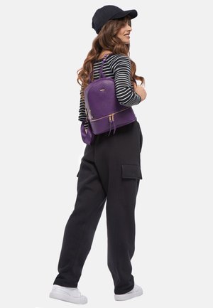 Zaino in pelle viola con hardware dorato, chiusura zip e doppie spalline; indossato con pantaloni cargo neri e una maglietta a righe.