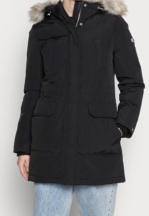 Manteau d'hiver noir à capuche avec bordure en fourrure, fermeture éclair et boutons, rabat avant et poches verticales, porté avec un jean bleu clair.