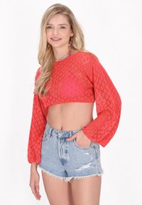 Horgolt stílusú, rövid piros pulóver hosszú, puffos ujjakkal és gyémánt mintázattal, magas derekú, kopott farmer sorttal párosítva.