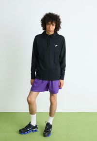 Črno dolg rokav hoodie z vrvicami in belim logotipom, v kombinaciji z vijoličnimi kratkimi hlačami, belimi nogavicami in črnimi supergami z modrimi poudarki.
