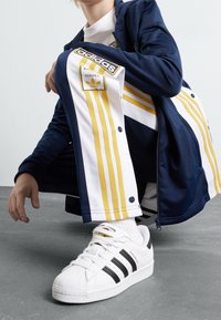 Námořnický a bílý sportovní oblek se žlutými pruhy, který má patentky a logo Adidas, doplněný o bílé tenisky Adidas Superstar.