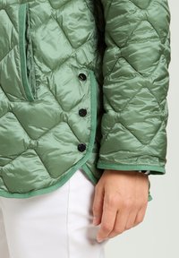Pikowana zielona kurtka puchowa z błyszczącą powierzchnią, z diagonalnym wzorem diamentowym, czarnymi zatrzaskami oraz kontrastującymi zielonymi wykończeniami.