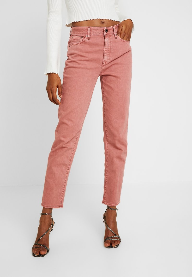 Kvinna i högmidjade rosa jeans, vit cropped långärmad topp och svarta strapless klacksandaler, poserar med handen i fickan.