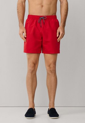 Uomo che indossa pantaloncini da nuoto rossi con cordoncini neri e bianchi e scarpe slip-on nere, in piedi contro uno sfondo chiaro e semplice.