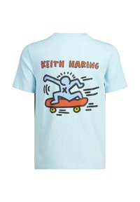 T-shirt di cotone azzurro chiaro con una grafica di una figura blu che fa skateboard su uno skateboard arancione con ruote gialle e la scritta "KEITH HARING".