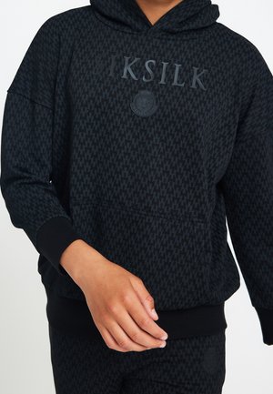 Sort tekstureret hættetrøje med "SIKSILK"-logo og løveemblem, båret med matchende sorte bukser, hånd placeret nær taljen.