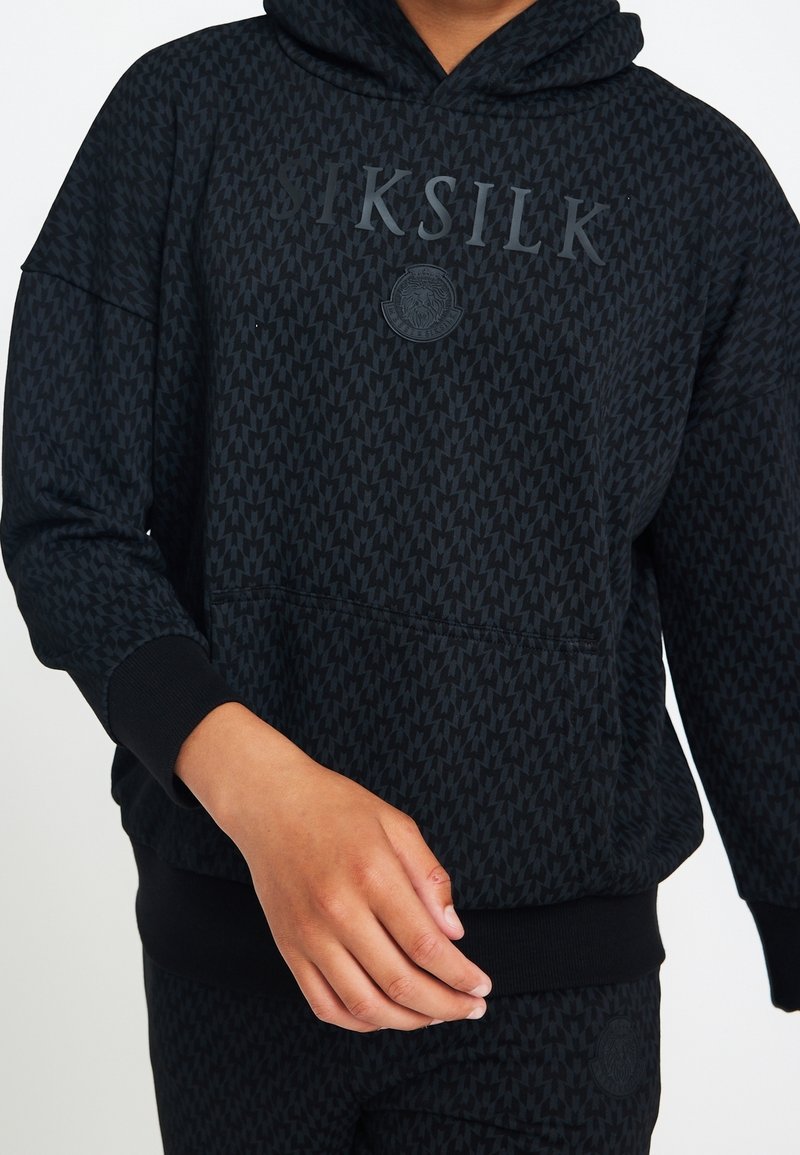 Sweat à capuche noir texturé avec logo "SIKSILK" et emblème de lion, porté avec un pantalon noir assorti, main positionnée près de la taille.