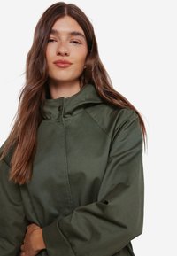 Olivengrøn jakke med høj krave, knapper og raglanærmer, lavet af et glat, holdbart stof. Ingen synlige mønstre eller hardware-detaljer.