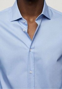 Persona di pelle scura che indossa una camicia azzurra chiaro con il primo bottone slacciato, mostrando parte del colletto e dell'area del petto.