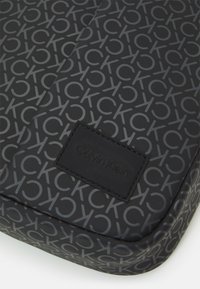 Borsa Calvin Klein con finitura nera opaca, caratterizzata da un motivo a logo grigio ripetuto e un'etichetta in pelle nera texturizzata.