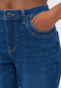 Modre denim hlače z visokim pasom, sprednjimi žepi in kovinskim gumbom za zapiranje. Imajo kontrastno šivanje in gladko teksturo.