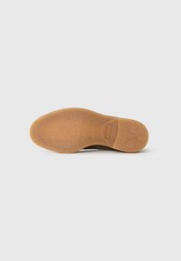 Suela de un zapato casual marrón con fondo de goma texturizada y un acento rectangular liso cerca del talón sobre un fondo blanco liso.