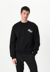 Sweatshirt preto com gola redonda, punhos com ribanas e um pequeno logotipo branco no lado esquerdo do peito. Textura suave e corte casual.